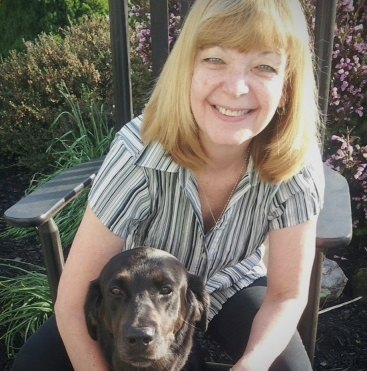 Woman with reddish-blonde hair smiling while holding a brown dog outdoors